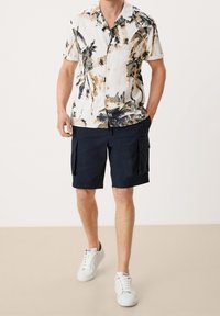 Camicetta a maniche corte con stampa tropicale di palme in beige e verde; abbinata a pantaloni cargo blu navy e sneakers bianche.