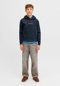 Sweat à capuche bleu marine avec poche avant et logo doré, superposé à une chemise bleue. Jean gris clair à jambes larges et chaussures marron, debout sur un fond neutre.