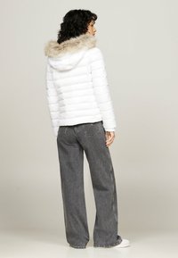 Giacca puffer bianca con cuciture orizzontali, cappuccio in pelliccia sintetica staccabile di colore beige, abbinata a jeans grigi larghi e sneakers bianche.