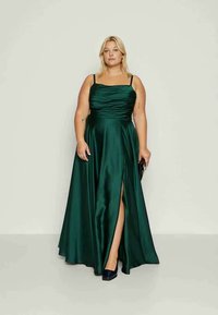 Smaragdin vihreä satiini-ilta-asu, jossa on istuva, röyhelöity bodice, ohuet hihnat ja reiden ylle ulottuva halkio, yhdistettynä clutch-laukkuun ja tummiin kenkiin.