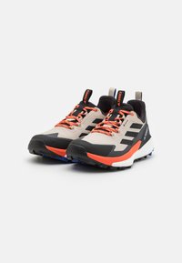 Men's trail running shoes with a beige mesh upper, black accents, and bright orange laces. Features a thick black sole with blue details.