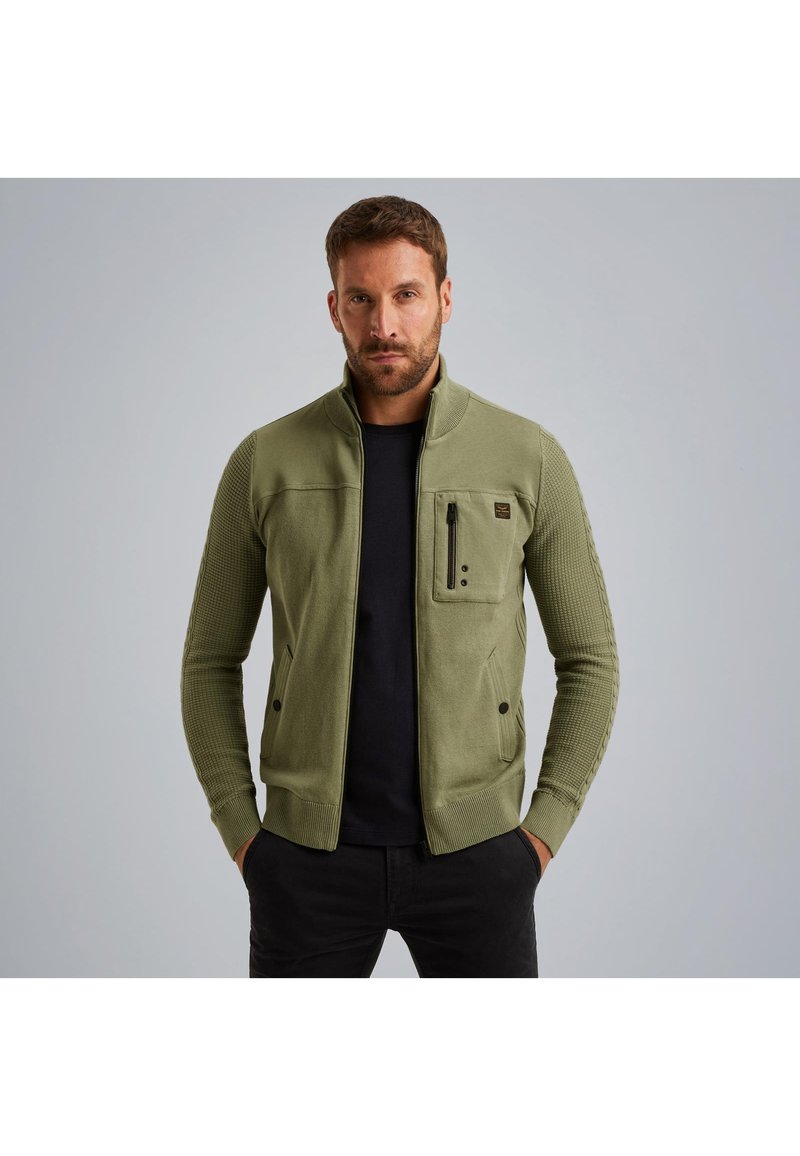 Olivno zeleni zip-up jopič z rebrasto teksturo, s stoječim ovratnikom in žepom na zadrgo. Ima prilegajočo obliko in stranske žepe.