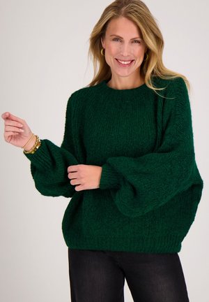 Vrouw glimlachend, gekleed in een donkergroen grofgebreid trui met ballonmouwen en zwarte broek, poseert met één hand vlak bij armbanden.