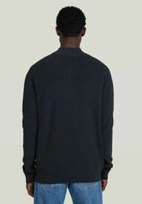 Donkergroene geribde trui met een ronde halslijn en lange mouwen, met een rechte zoom en een gestructureerde stof. Draagt over lichtblauwe jeans.