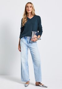 Donker teal v-hals trui, lichtblauwe wijde jeans en zilveren clutch. Platte schoenen met glanzende puntneuzen. Eenvoudig, modern ontwerp.