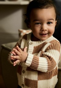 Der gestrickte Cardigan verfügt über abwechselnde braune und cremefarbene Blöcke, mit einem gerippten Kragen, Bündchen und einer Knopfleiste. Die Hände sind vorne gefaltet.