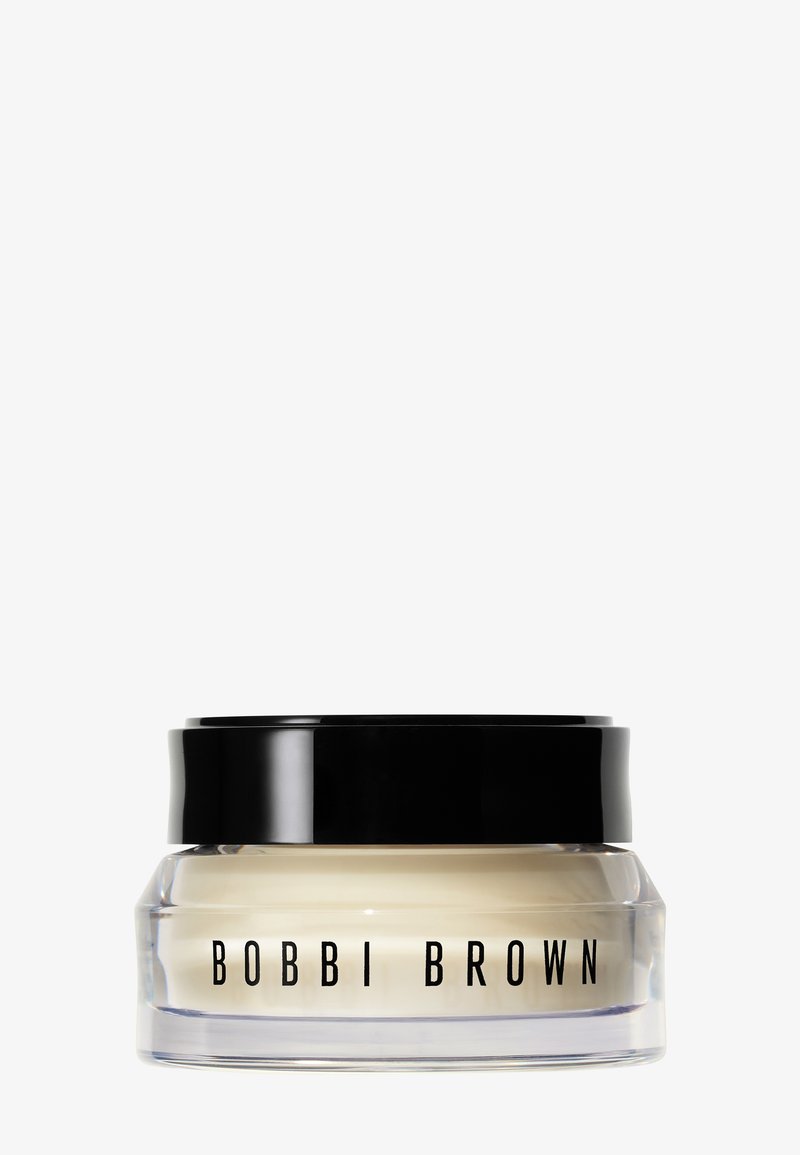 Pot en verre transparent avec un couvercle noir contenant une crème étiquetée Bobbi Brown sur un fond clair uni.