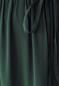 Green textured fabric with vertical pleats and a tied waist. The material appears lightweight and flowy, enhancing its draping quality.