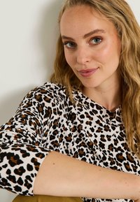 Top de estampado de leopardo con base blanca, mangas largas, textura acanalada y acentos marrones. Primer plano de la tela y detalles de diseño.