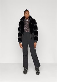 Casaco bomber de pelo sintético preto com textura riscada, usado sobre uma blusa padronizada e combinado com calças pretas e botas de esqui.