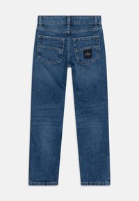 Denimjeans i mellanmörkblå färg med rak skärning, två bakfickor och en svart logotypmärke på högerfickan. Slät textur.