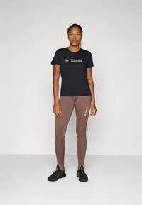 Ženska stoji v črni majici Terrex, rjavih legicah Terrex in črnih športnih čevljih proti enostavni beli ozadju.