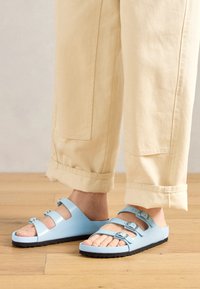 Light blue slide sandals with three adjustable straps and silver buckles; textured sole and smooth upper material. Worn with rolled-up beige pants.