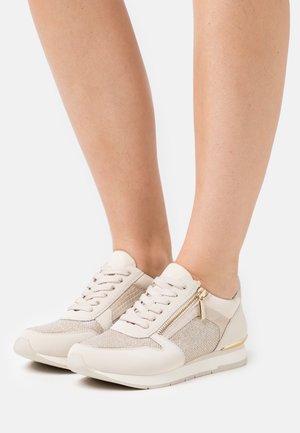 Beige tenisky s texturovanými látkovými panely, plochými tkaničkami a metalickými detaily. Boční zip pro snadný přístup. Gumová podešev.