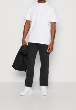 Camiseta blanca de manga corta con el logotipo de "Calvin Klein Jeans" en el cuello, combinada con unos vaqueros negros de pierna recta y zapatillas blancas. Chaqueta negra en la mano.