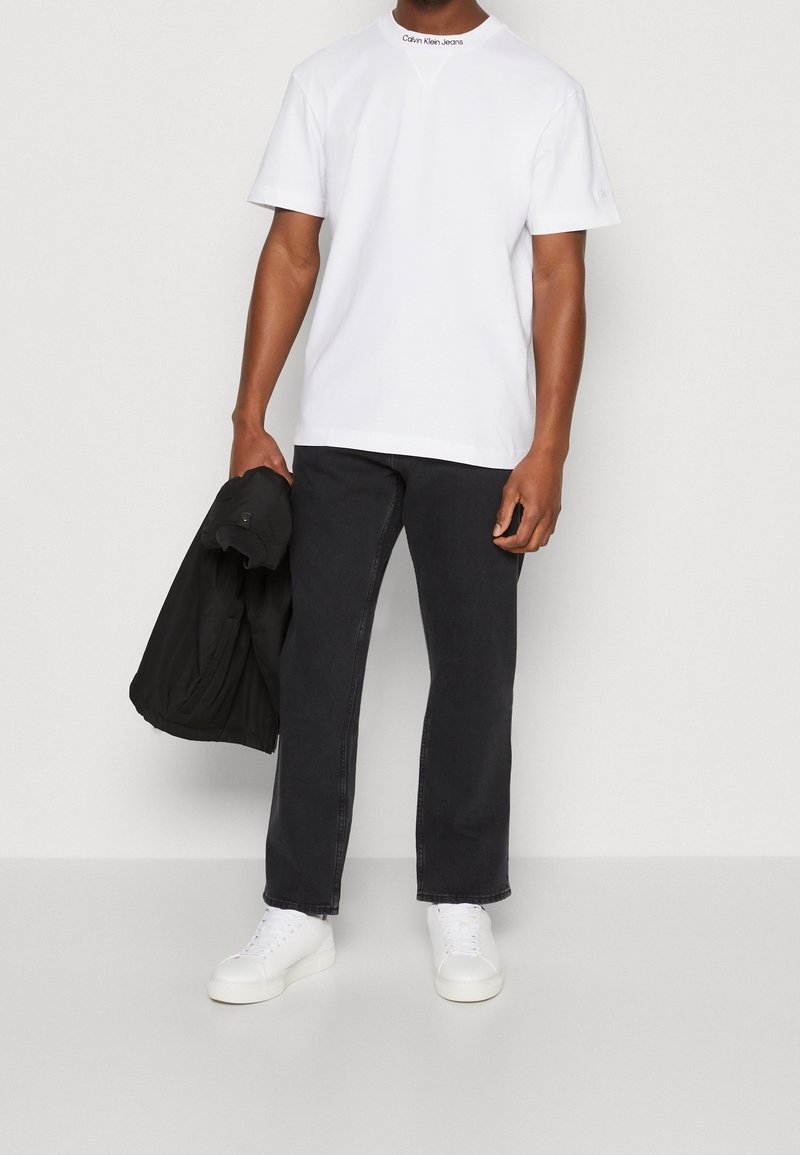 Camiseta blanca de manga corta con el logotipo de "Calvin Klein Jeans" en el cuello, combinada con unos vaqueros negros de pierna recta y zapatillas blancas. Chaqueta negra en la mano.