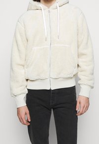 Cremefarvet zip-hoodie lavet af fluffy materiale, med hætte, forlommer og hvide snore. Ribbet ved håndleddene og kanten.