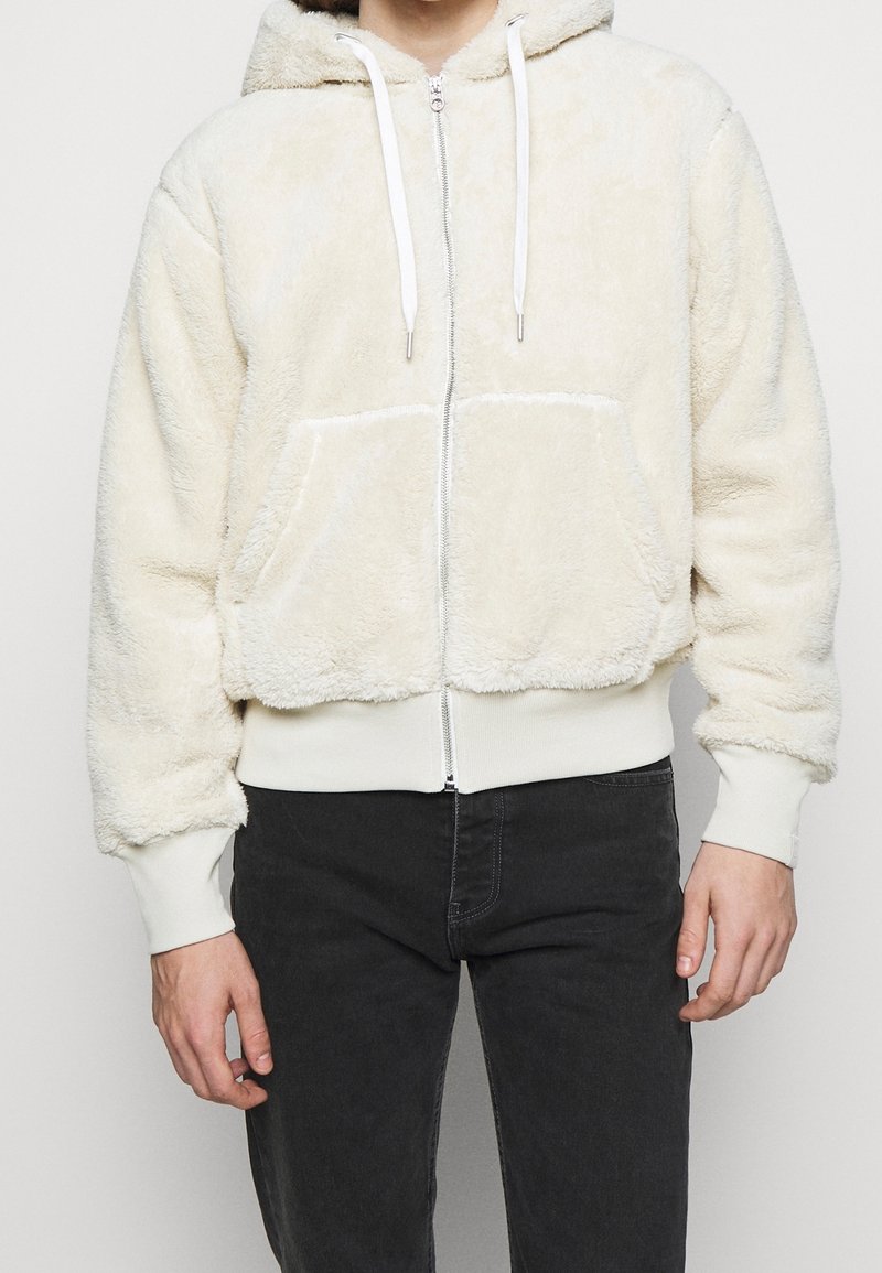 Cremefarvet zip-hoodie lavet af fluffy materiale, med hætte, forlommer og hvide snore. Ribbet ved håndleddene og kanten.