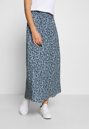 Femme portant une jupe midi fleurie bleue avec une ceinture élastique, des baskets blanches et un haut blanc, debout devant un fond uni.