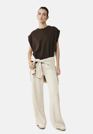 Vrouw draagt donkerbruine mouwloze top en crèmekleurige wijde broek met een licht jasje om haar taille gebonden, staand tegen een witte achtergrond.