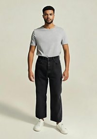 Bărbat stând cu fața înainte, purtând un tricou cu mânecă scurtă gri deschis, blugi negri cu croială largă și adidași albi pe un fundal simplu.