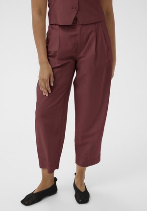 Donna che indossa pantaloni corti color bordeaux e scarpe slip-on nere in rete, in piedi con una mano appoggiata sulla coscia su sfondo semplice.