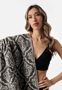 Coperta decorativa grigia con intricati motivi damascati bianchi, texture morbida e bordo chiaro a contrasto, tenuta da un modello.