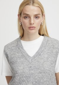 Grauer, strukturierter Sweater-Weste mit V-Ausschnitt, die über einem weißen T-Shirt getragen wird. Die Weste hat ein zartes Muster und ist aus weichem, gestricktem Stoff gefertigt.