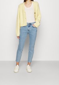 Cardigan côtelé jaune avec fermeture à boutons, porté sur un haut clair et un jean taille haute bleu, associé à des baskets blanches.