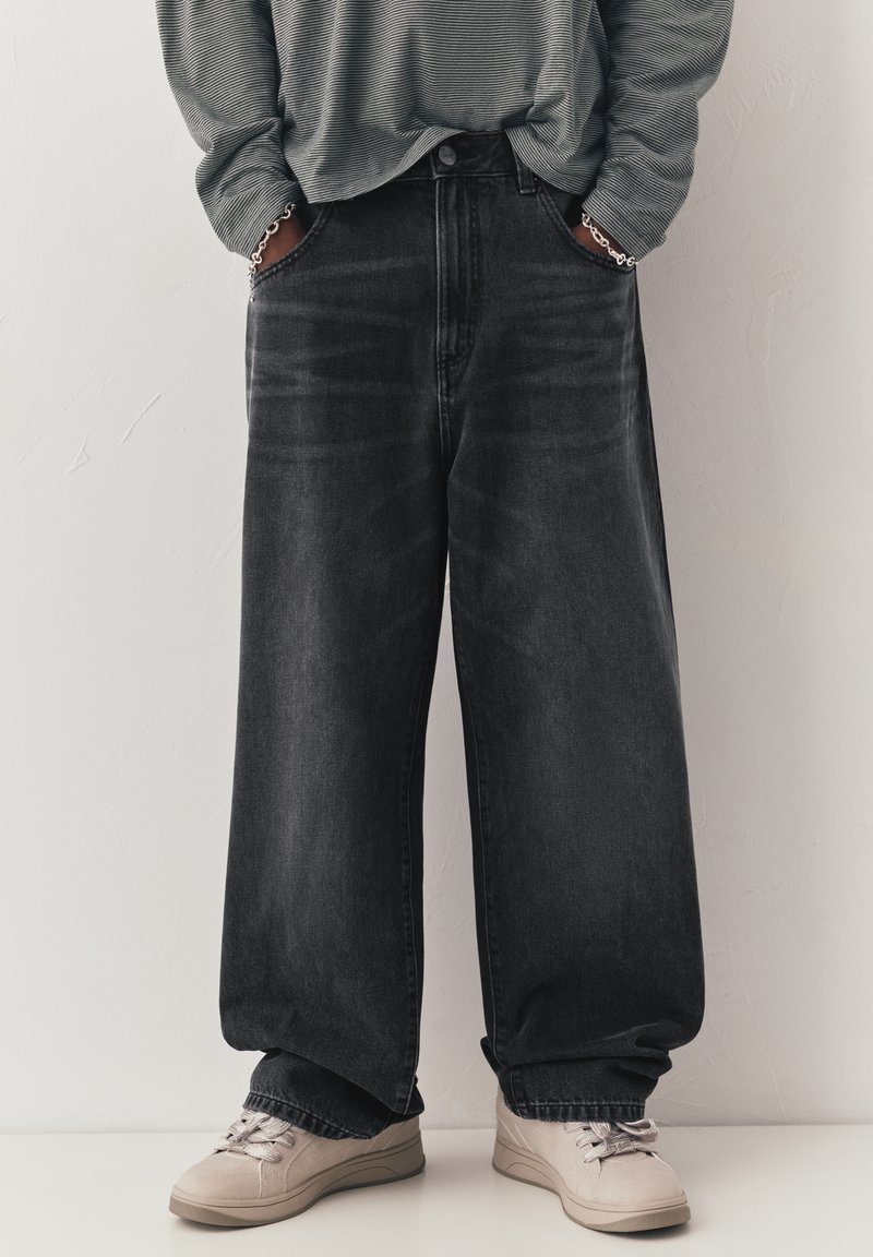 Personne portant un jean noir à jambes larges avec les mains dans les poches, un pull à manches longues rayé, des bracelets argentés et des baskets beige clair.