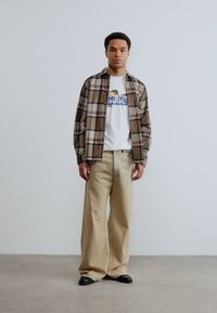 Άνδρας που στέκεται φορώντας μπεζ παντελόνι με φαρδιά πόδια, μαύρα παπούτσια, λευκό μπλουζάκι με στάμπα και καφέ πουκάμισο με καρό κουμπιά, μπροστά σε απλό φόντο.