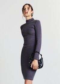 Figurbetontes, langärmeliges Kleid aus dunkelviolettem Stoff mit hohem Ausschnitt. Modell hält eine kleine schwarze Tasche mit silbernen Akzenten.