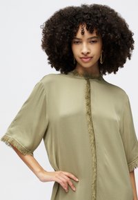 Olivová blúzka s krátkymi rukávmi, s fr-kaním lemem okolo krku a rukávov. Hladká, ľahká tkanina s voľným strihom.