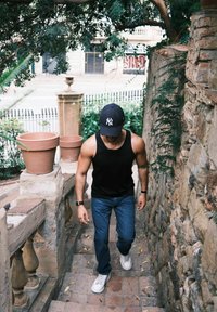 Mann iført svart ermeløs topp, jeans og hvite sko som går opp utendørs steintrapper mellom steinvegger med potteplanter i nærheten.