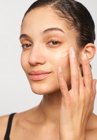 Femme appliquant une crème beige clair sur sa joue avec ses doigts, affichant une peau lisse, un maquillage naturel et des cheveux courts et mouillés.