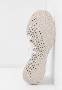 Semelle de basket beige et blanche avec des motifs de grip hexagonaux et circulaires appuyée contre un mur blanc.