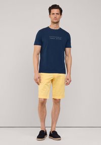 Camiseta de manga corta azul marino con texto blanco, combinada con pantalones cortos amarillos. El modelo lleva zapatos negros tipo slip-on. Diseño simple y casual.