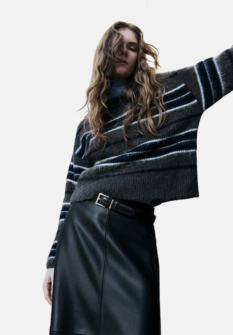 Donna con capelli mossi che indossa un maglione a righe grigio scuro e una gonna di pelle nera con cintura, in posa davanti a uno sfondo neutro.