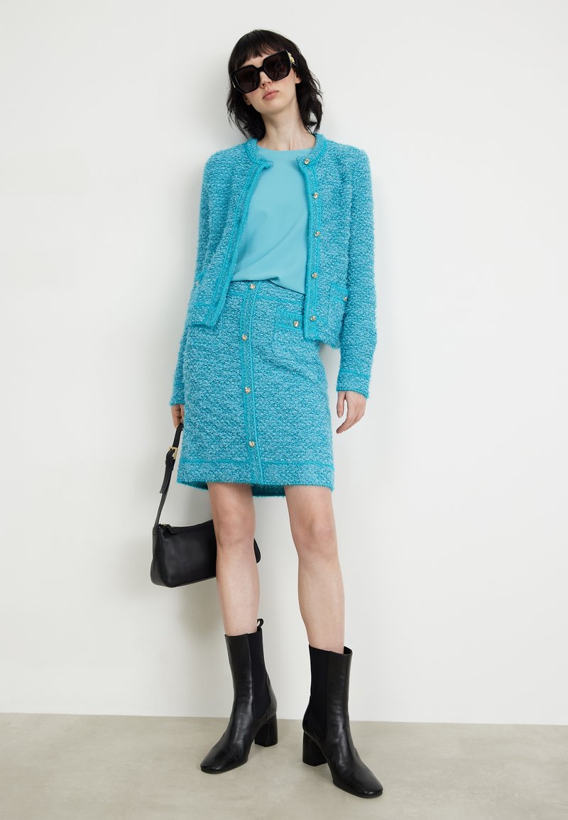 Türkises Zwei-Teiler-Outfit bestehend aus einer strukturierten, knöpfbaren Strickjacke und einem passenden Rock. Schwarze Knöchelstiefel und eine kleine schwarze Handtasche.