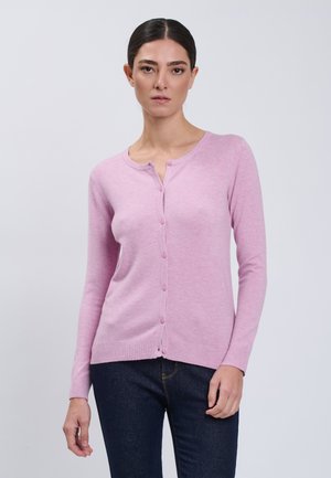 Frau trägt eine hellviolette, geknöpfte Strickjacke und dunkelblaue Jeans und steht vor einem schlichten hellgrauen Hintergrund.