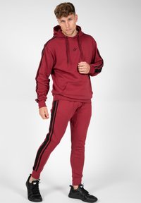 Bordeaux sweatshirt met zwarte zijkanten, capuchon en voorzak, gecombineerd met bijpassende bordeaux joggers en zwarte sport schoenen.