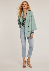 Blazer doppiopetto verde menta con grandi bottoni neri, indossato sopra una blusa nera a fiori. Combinato con jeans cropped blu chiaro e décolleté con cinturini.