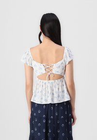 Blusa bianca floreale con maniche corte, scollatura sul retro e dettaglio con lacci. Il tessuto testurizzato e l'orlo arricciato aggiungono volume. Abbinata a una gonna navy con motivo.