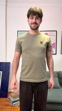 Νεαρός άνδρας με καστανά μαλλιά και γενειάδα, φορώντας ελιάς πράσινο μπλουζάκι και μαύρο παντελόνι, στέκεται σε ένα σαλόνι με καδραρισμένα έργα τέχνης και στοιβαγμένα βιβλία κοντά του.