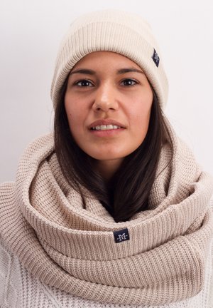 Beige knit beanie and textured scarf, both made of soft material. Scarf has a folded design with a small logo patch. Light background.