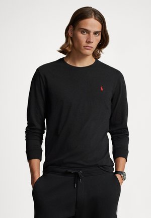 Black long-sleeve t-shirt, cotton material, ribbed cuffs, round neckline, features small red logo on left chest. Model wears black pants.