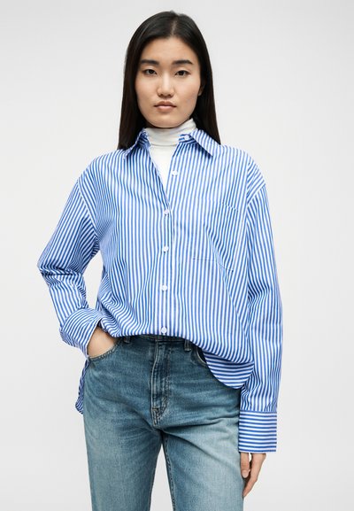 Νέα γυναίκα που φοράει μπλε και λευκό πουκάμισο με κάθετες ρίγες πάνω από λευκό ζιβάγκο και ανοιχτόχρωμο τζιν, ποζάροντας με το ένα χέρι στην τσέπη.