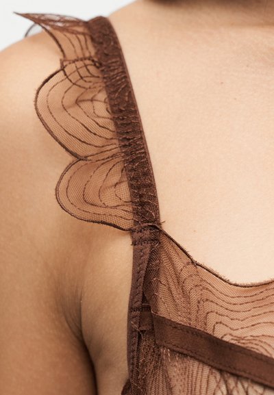Gros plan d'une épaule portant une bretelle en tissu marron transparent avec un détail en dentelle ondulée.