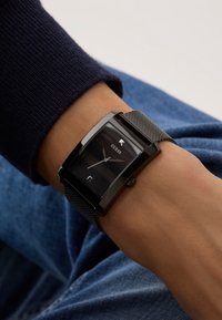 Schwarze rechteckige Uhr mit einem Mesh-Armband, ausgestattet mit einem dunklen Ziffernblatt, silbernen Zeigern und einem Datumsfenster. Metallgehäuse und polierte Akzente.