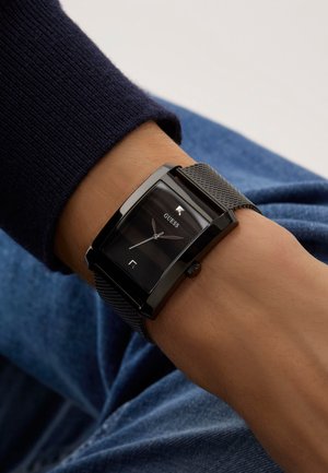 Montre rectangulaire noire avec un bracelet en maille, affichant un cadran sombre, des aiguilles argentées et une fenêtre de date. Boîtier en métal et accents polis.